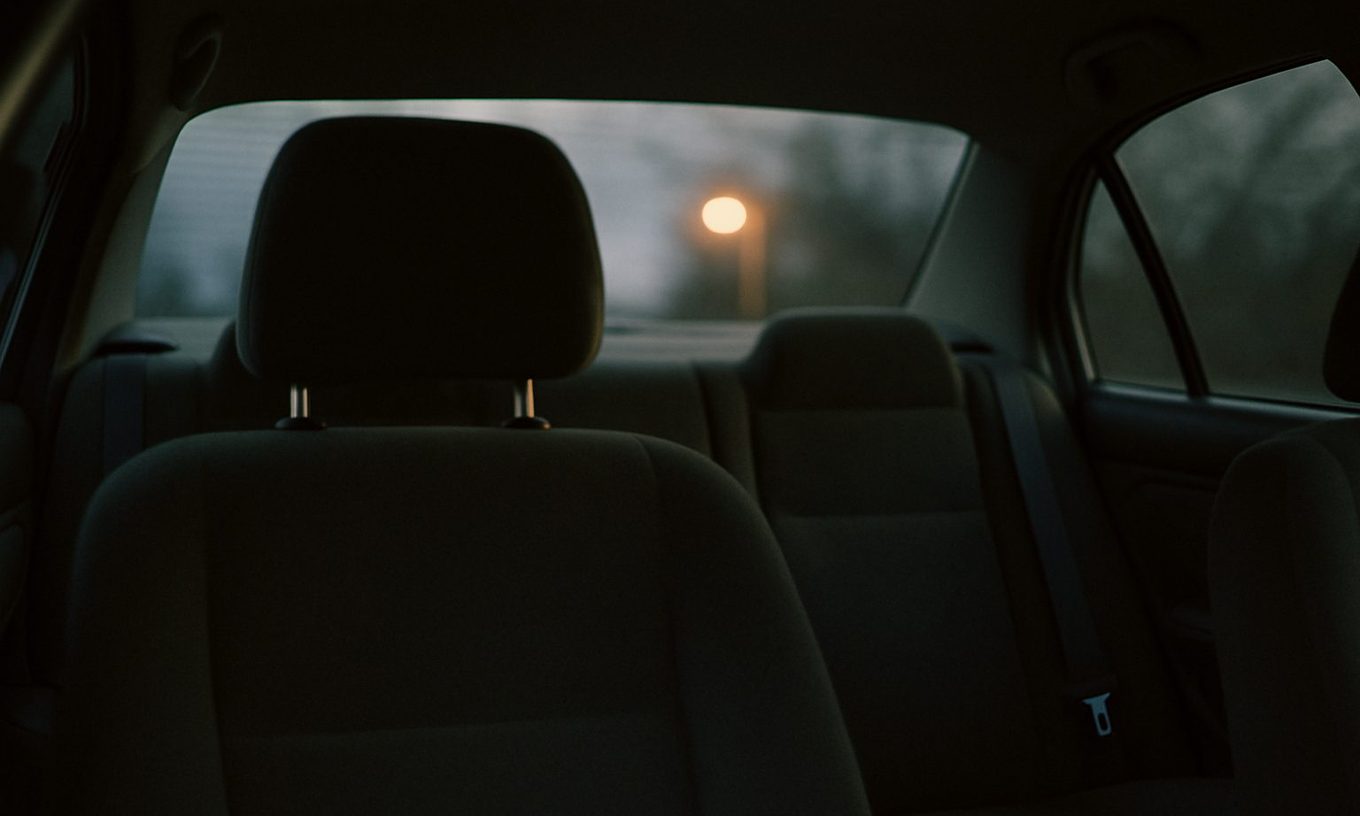 The inside of a parked sedan at dusk seen from the back seat, the driver's headrest in shadow and a single streetlight glowing through the rear window